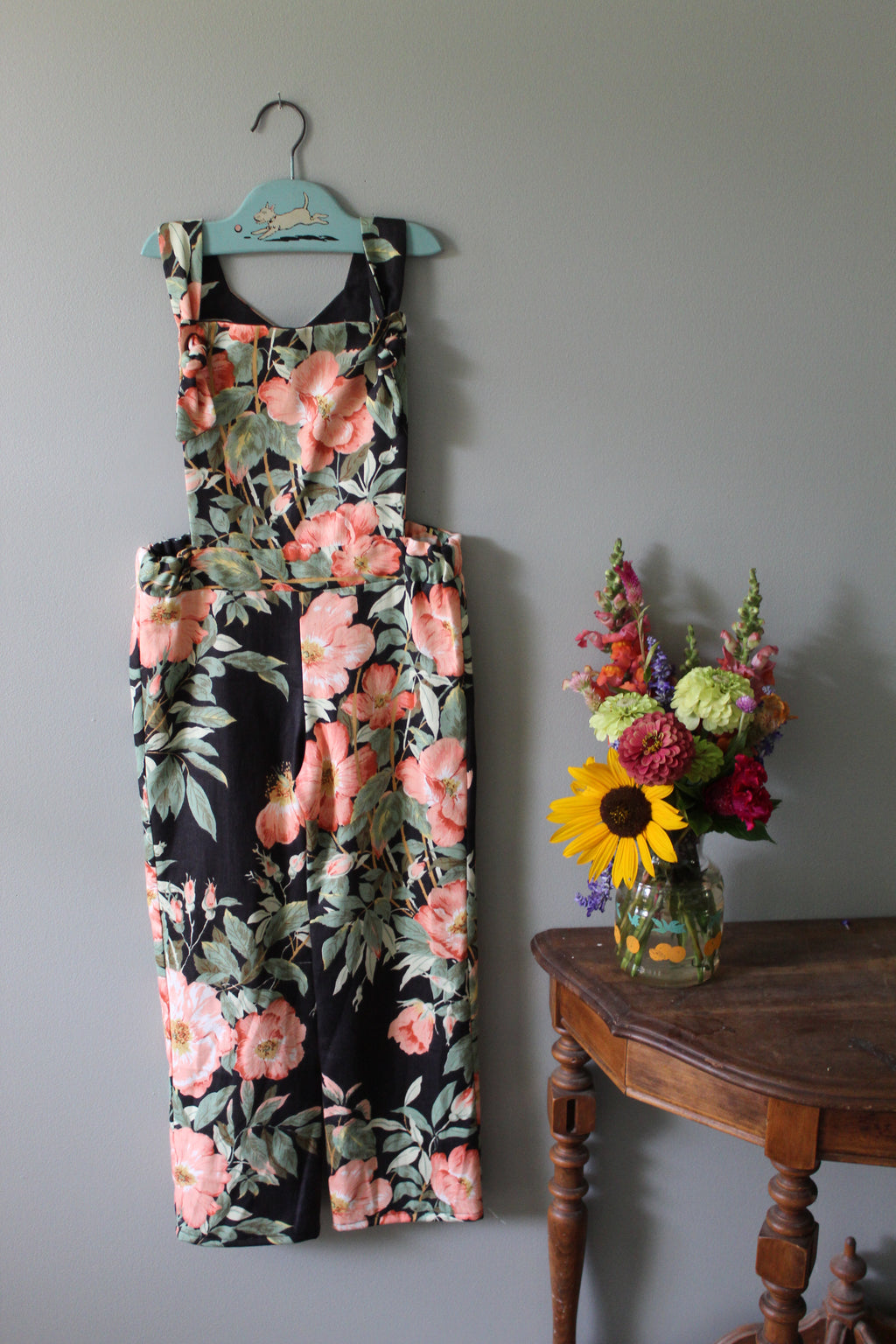 Floral-patterned overalls on a hanger with a small floral arrangement below on a gray wall.