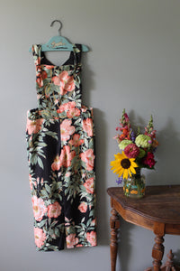 Floral-patterned overalls on a hanger with a small floral arrangement below on a gray wall.