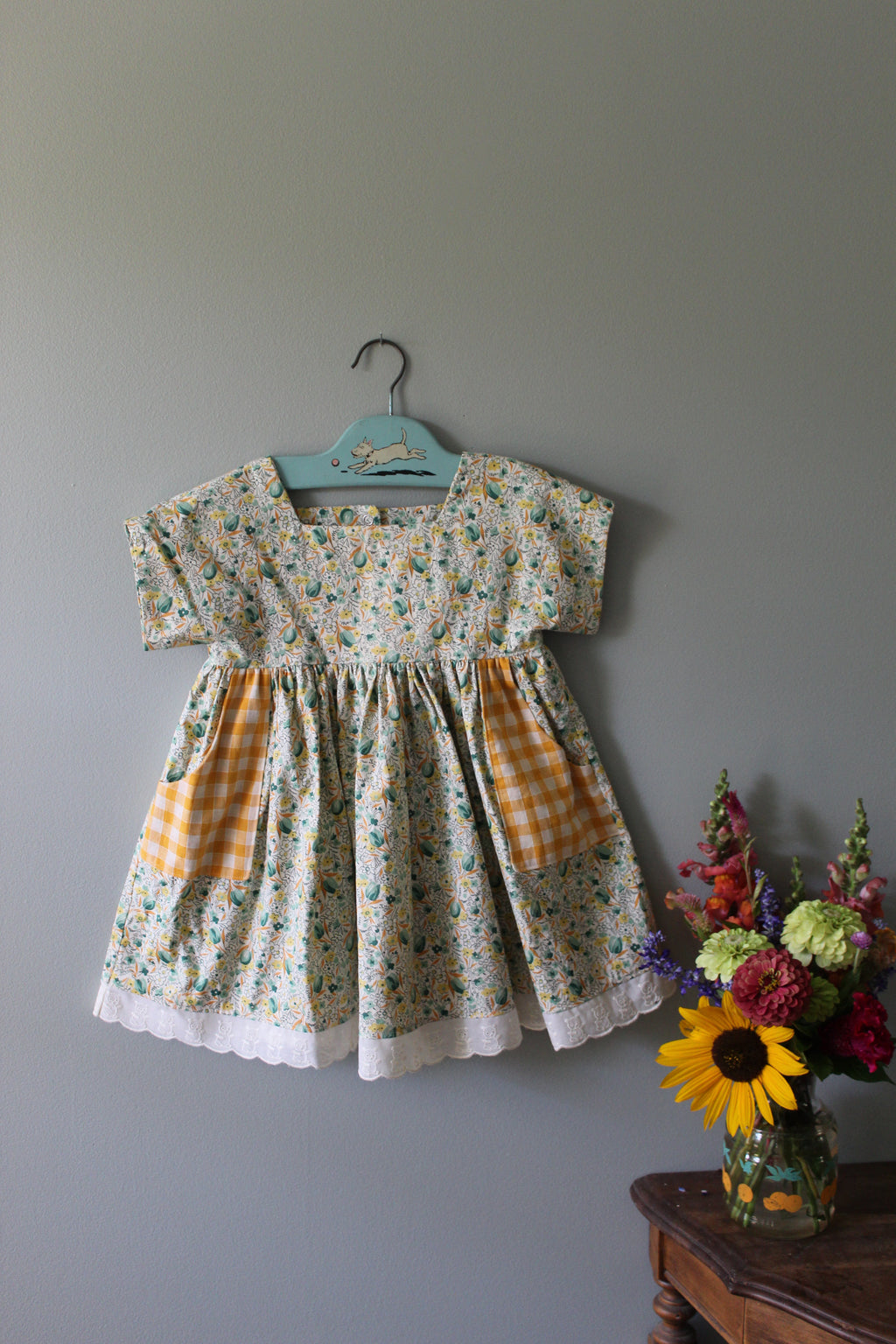 Floral tunic with checkered pocket on a hanger against a gray wall with flowers.