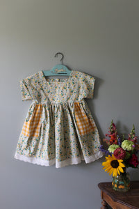 Floral tunic with checkered pocket on a hanger against a gray wall with flowers.