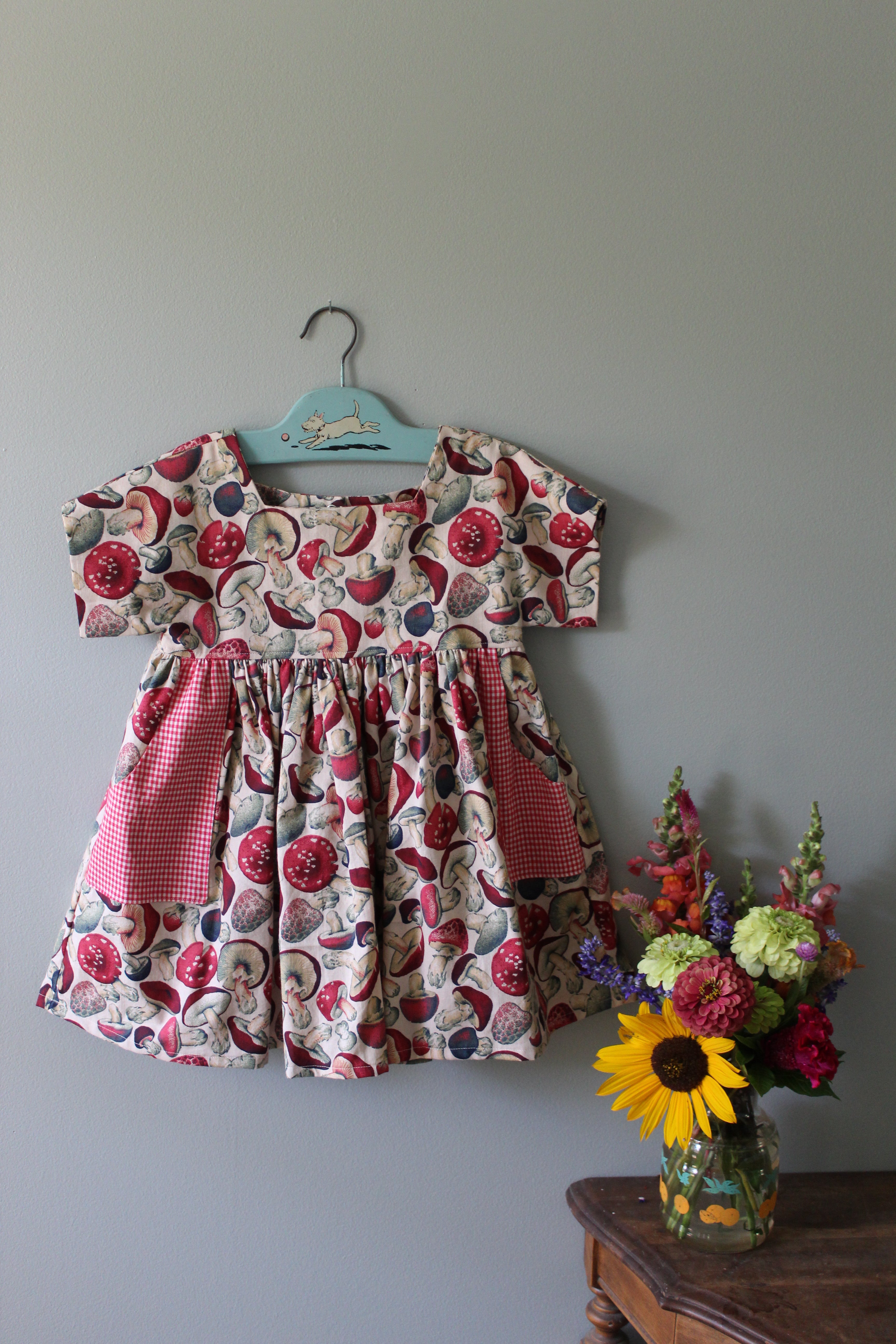 Floral-patterned girls tunic on a hanger with a small bouquet of flowers on a gray surface.