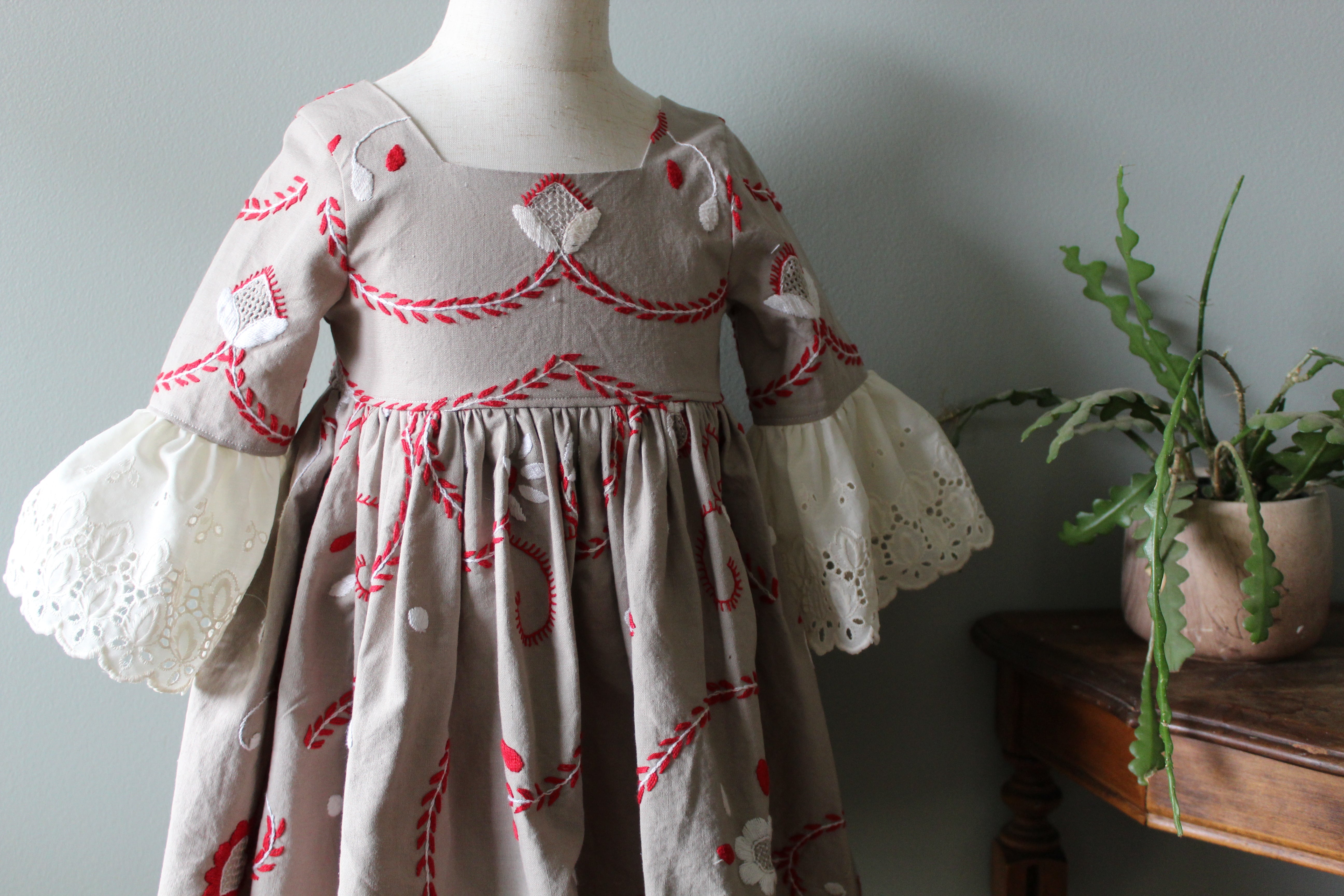 Children's dress with red embroidery on a mannequin against a plain wall.