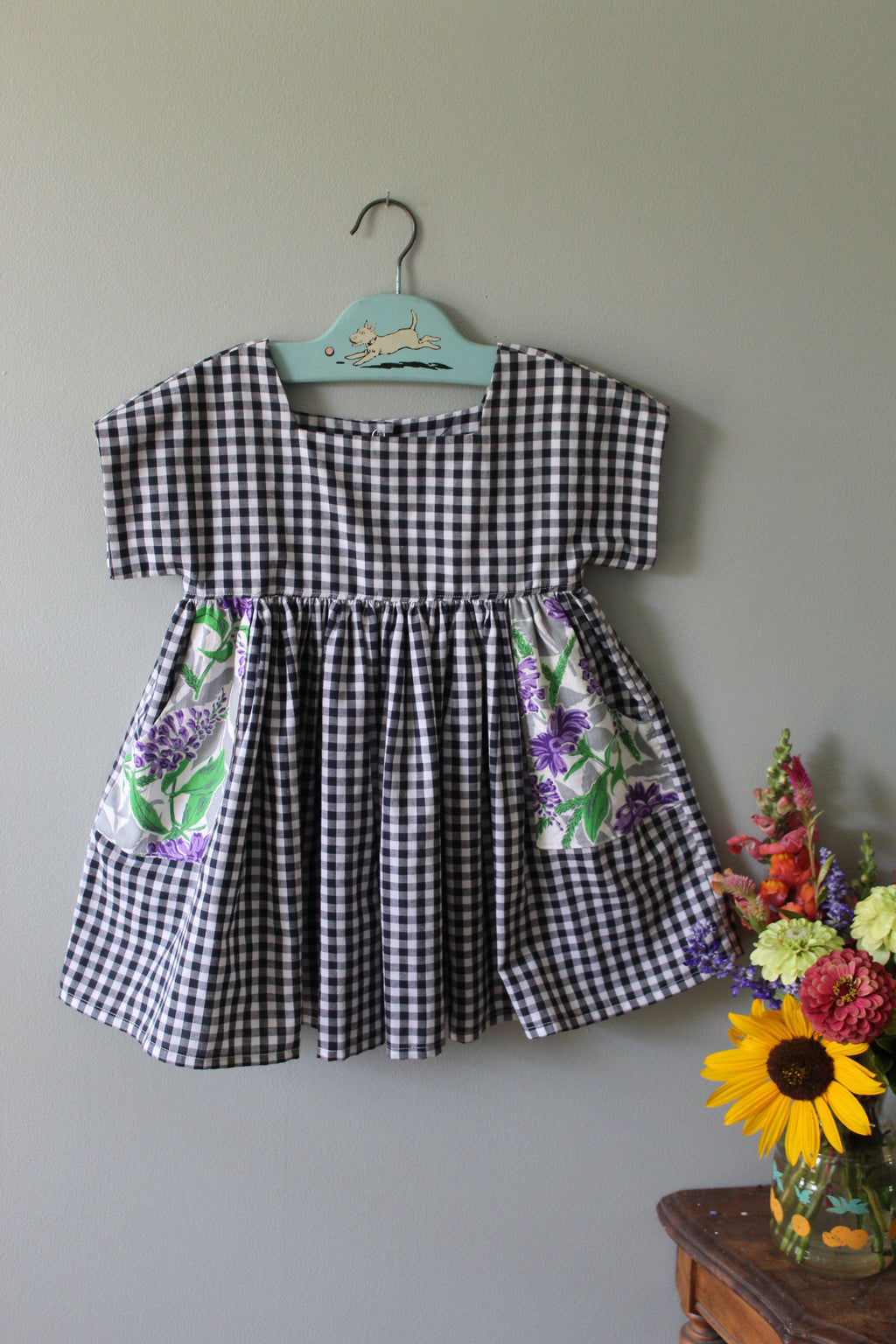 Black and white checkered tunic with floral accents on a hanger against a gray background