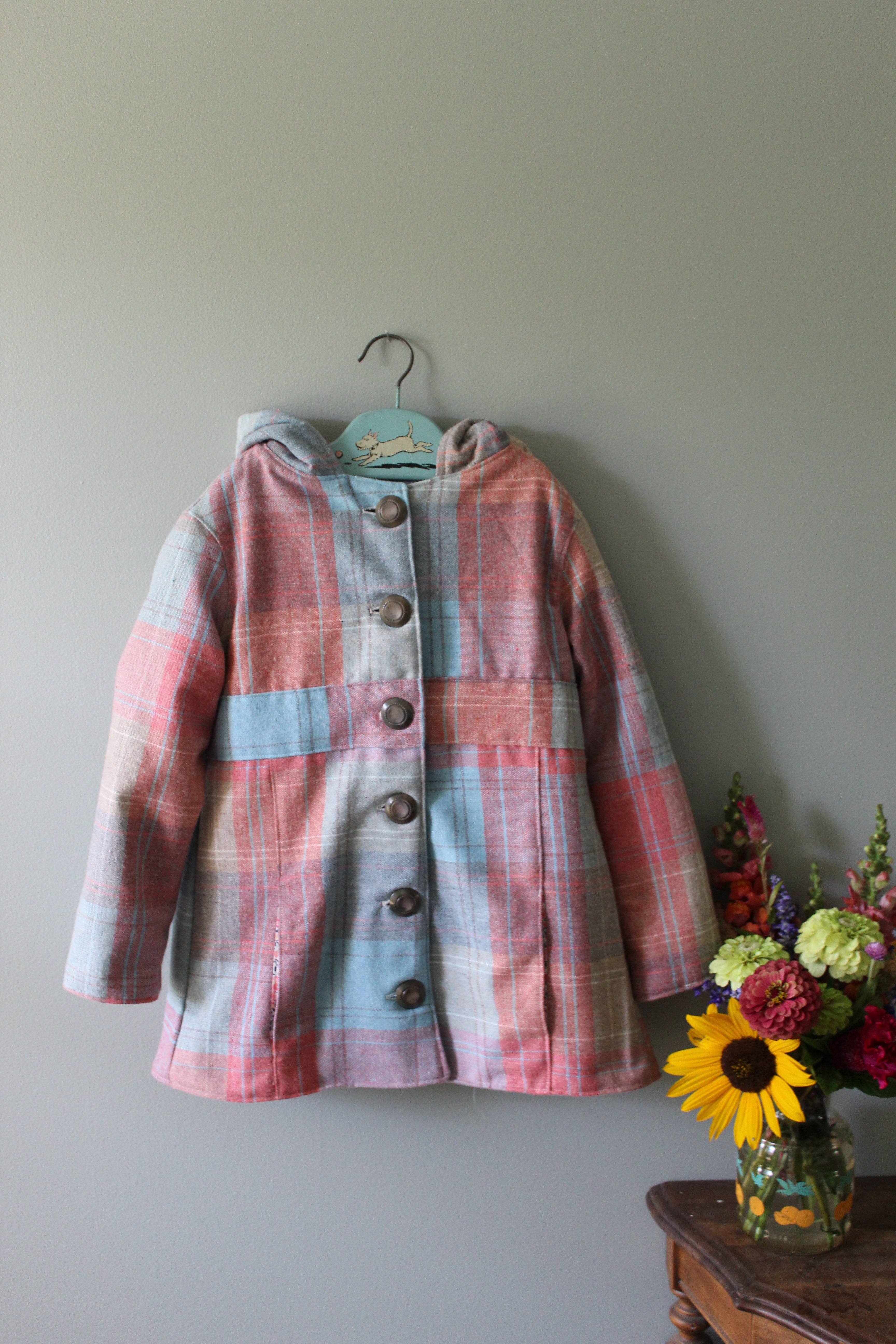 Pink plaid jacket on a hanger against a light green wall with flowers in the foreground.
