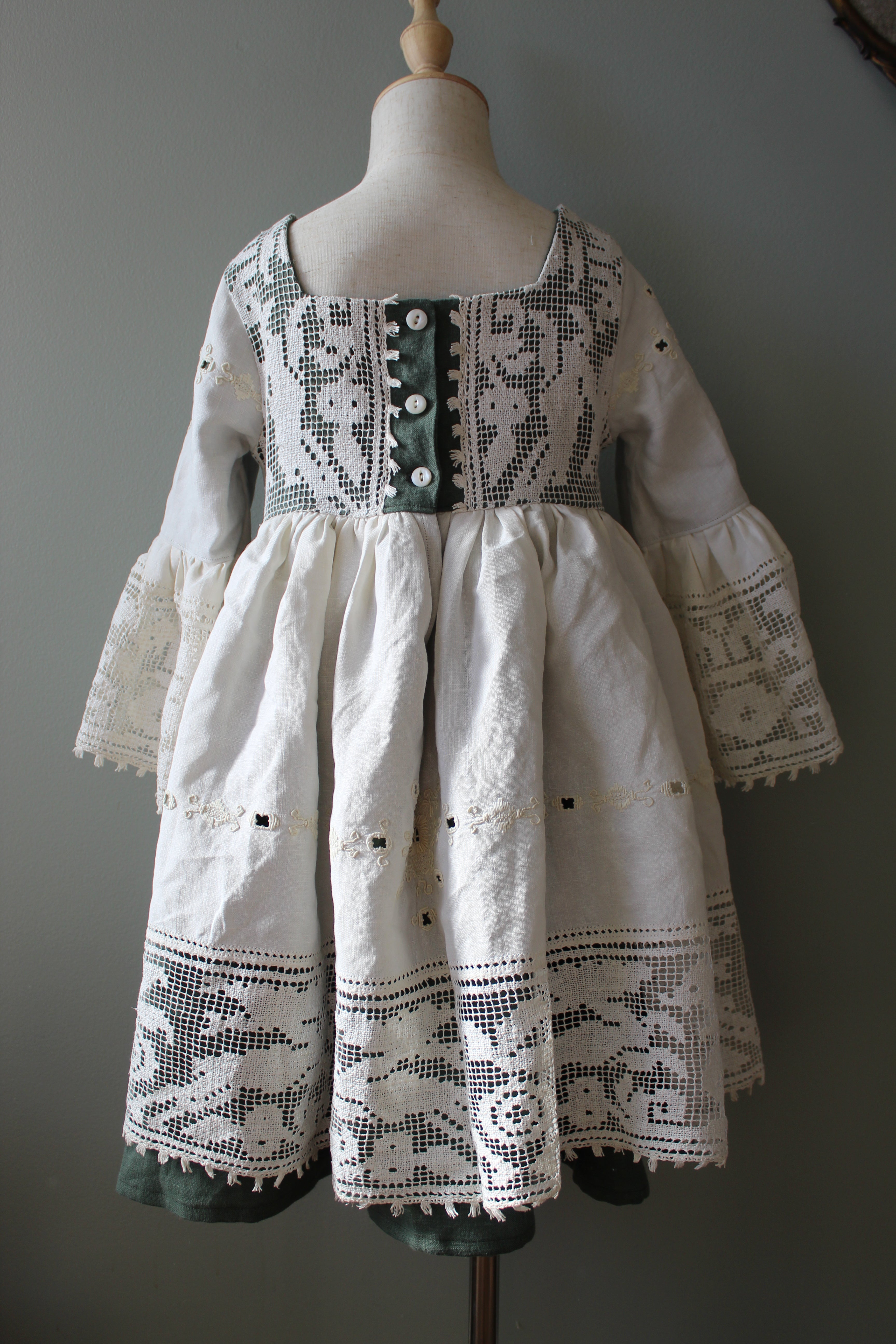 Mannequin wearing a white dress with intricate green lace details on a plain background