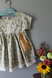 Floral tunic with checkered bottom section on a hanger next to a bouquet of flowers.