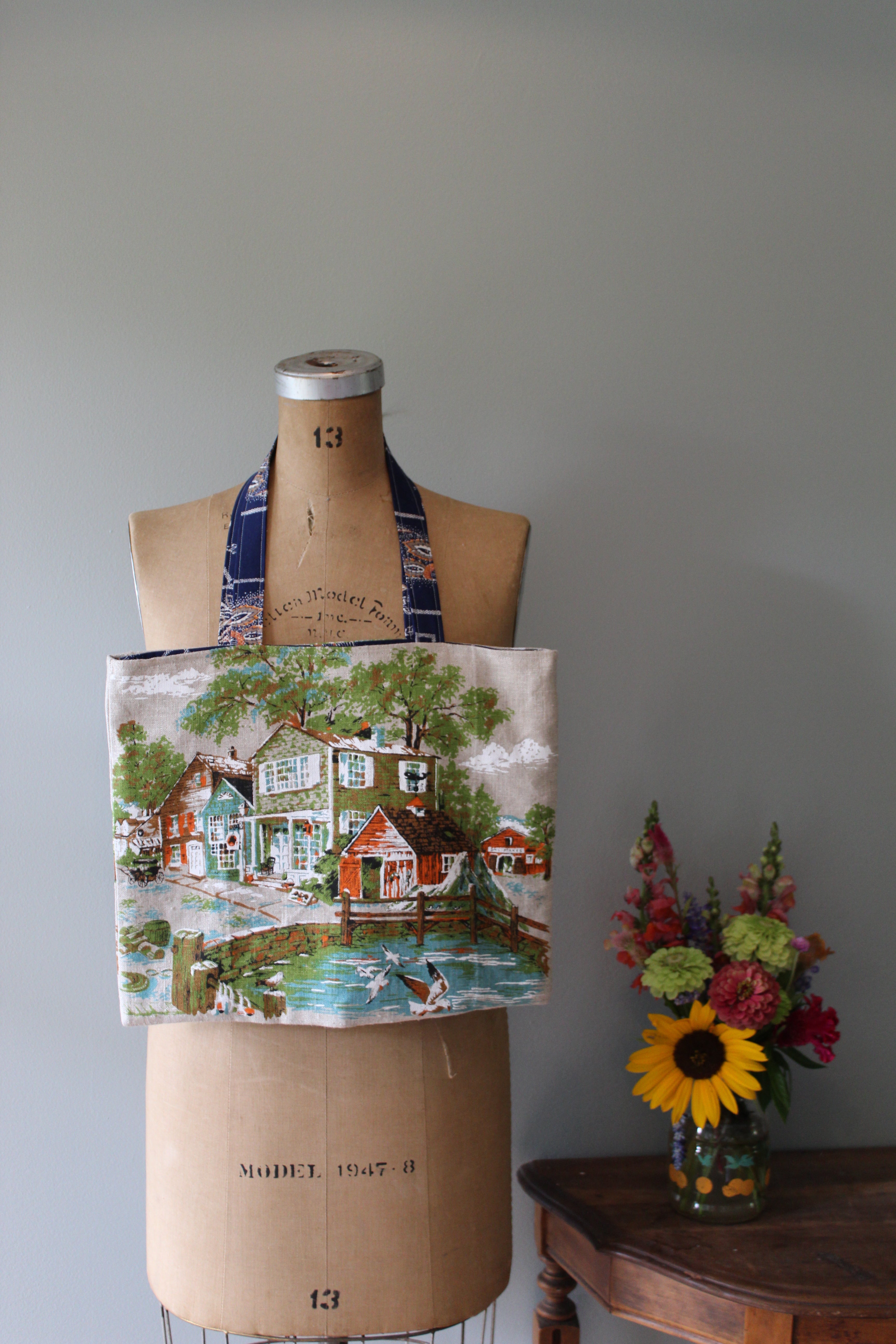 Mannequin wearing a patterned fabric tote bag with a scenic design, next to a vase of flowers on a wooden surface.