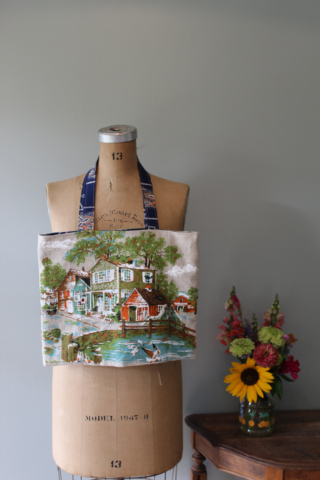 Mannequin wearing a patterned fabric tote bag with a scenic design, next to a vase of flowers on a wooden surface.