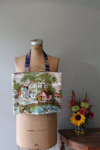 Mannequin wearing a patterned fabric tote bag with a scenic design, next to a vase of flowers on a wooden surface.