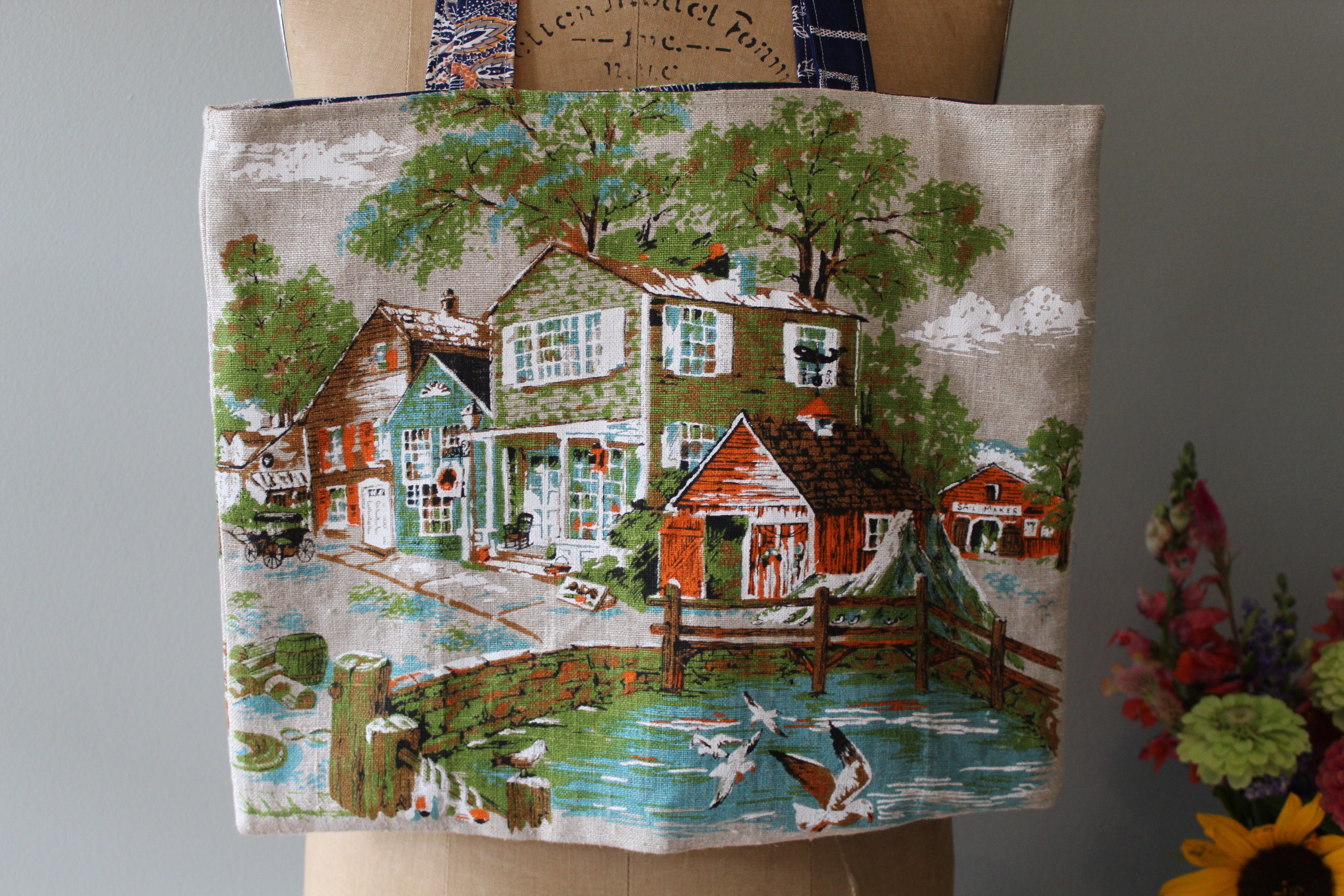 Tote bag with a printed design of a village scene held by a person.