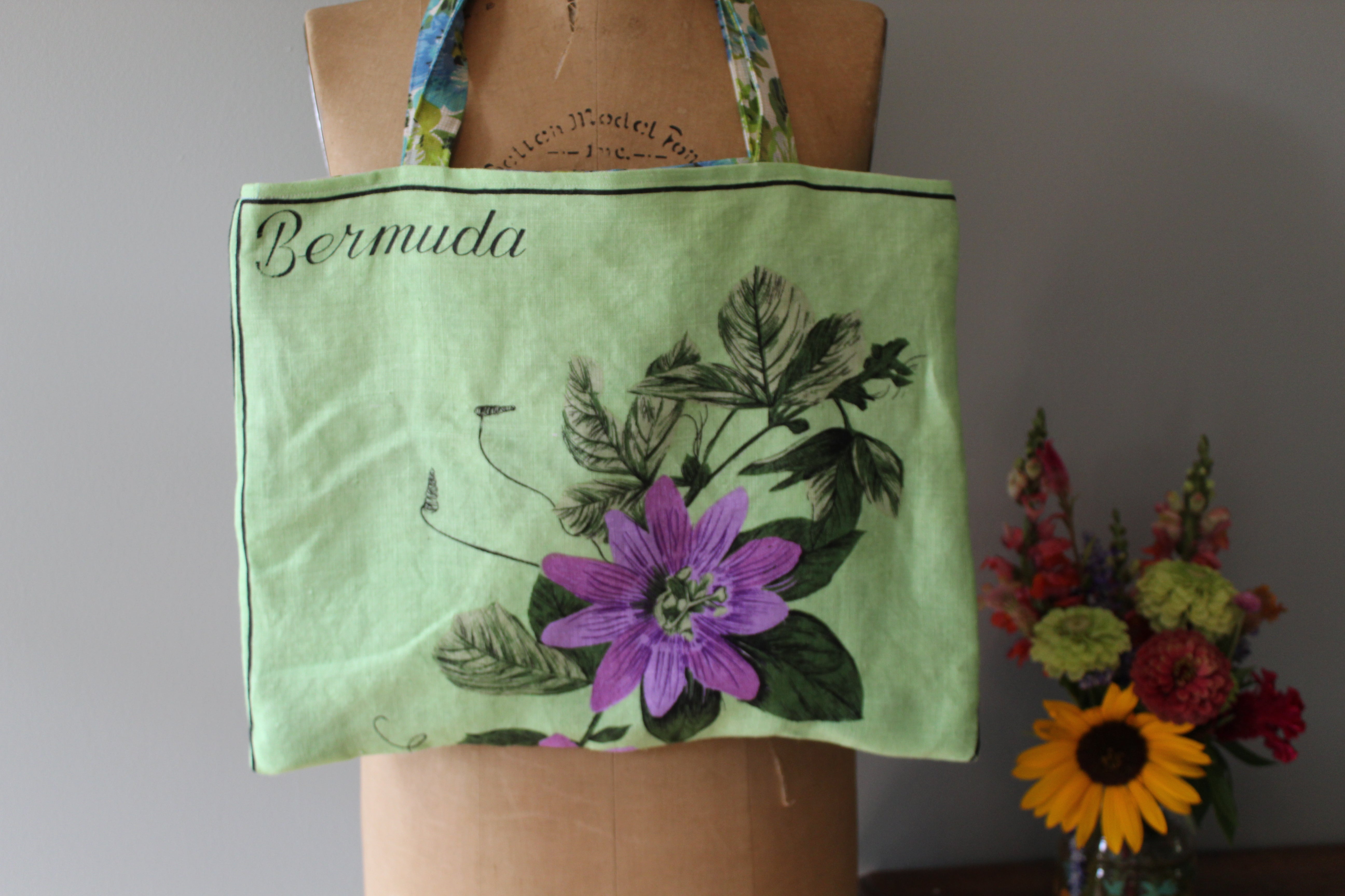 Green tote bag with floral design and 'Bermuda' text on a mannequin, with flowers in the background.