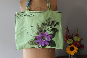 Green tote bag with floral design and 'Bermuda' text on a mannequin, with flowers in the background.
