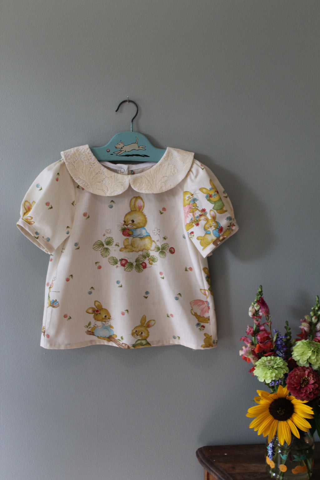 Children's top with bunny pattern on a hanger against a gray wall with flowers in the corner.