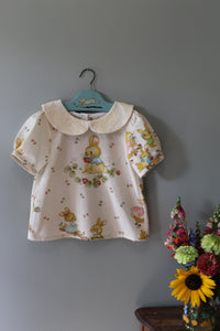 Children's top with bunny pattern on a hanger against a gray wall with flowers in the corner.
