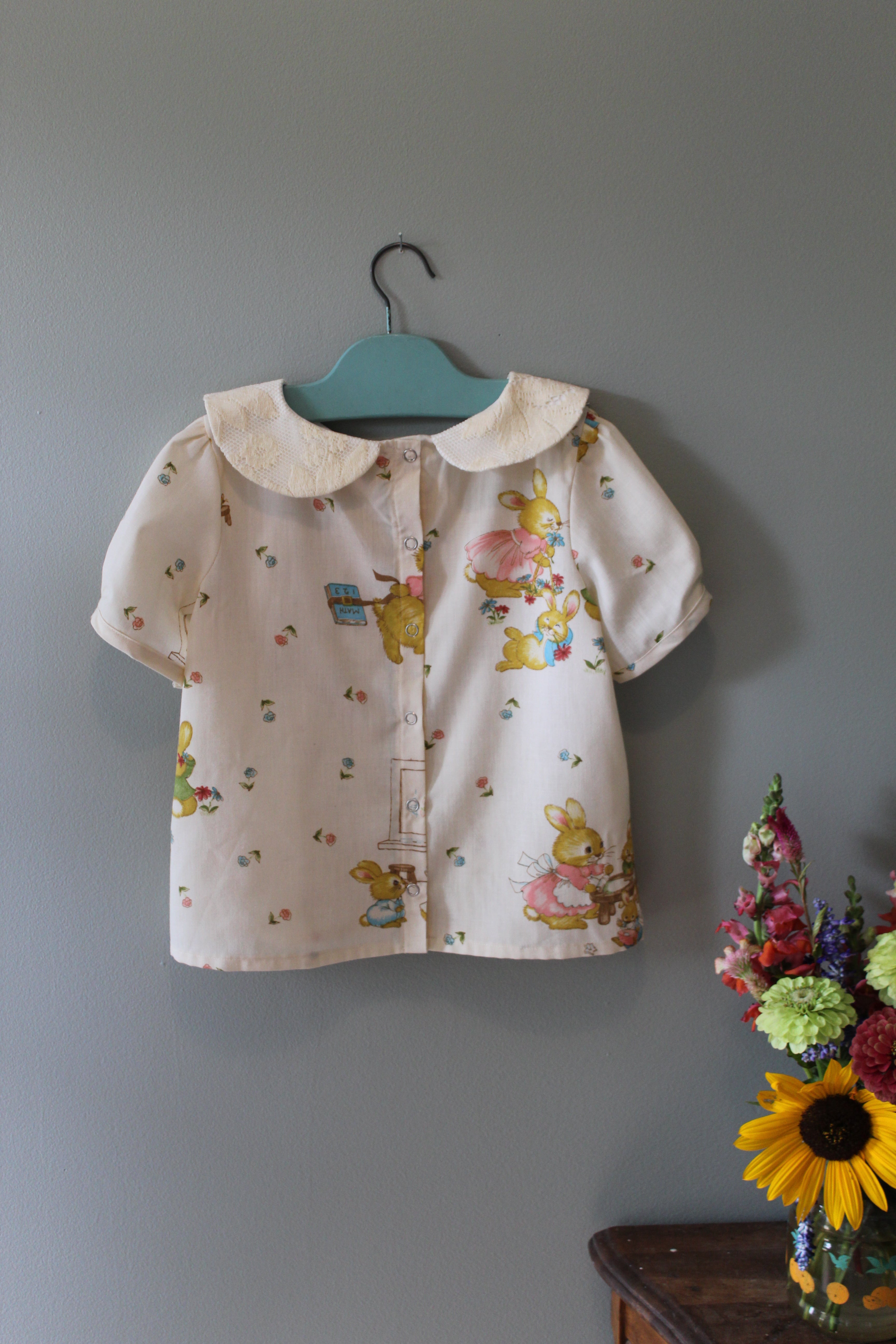 Children's top with bunny characters on a hanger against a gray wall, with flowers in the foreground.