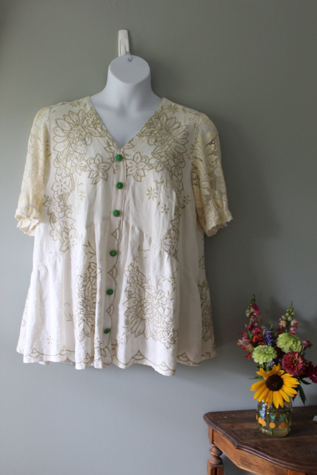 White lace top on a mannequin against a gray wall with flowers in the foreground.