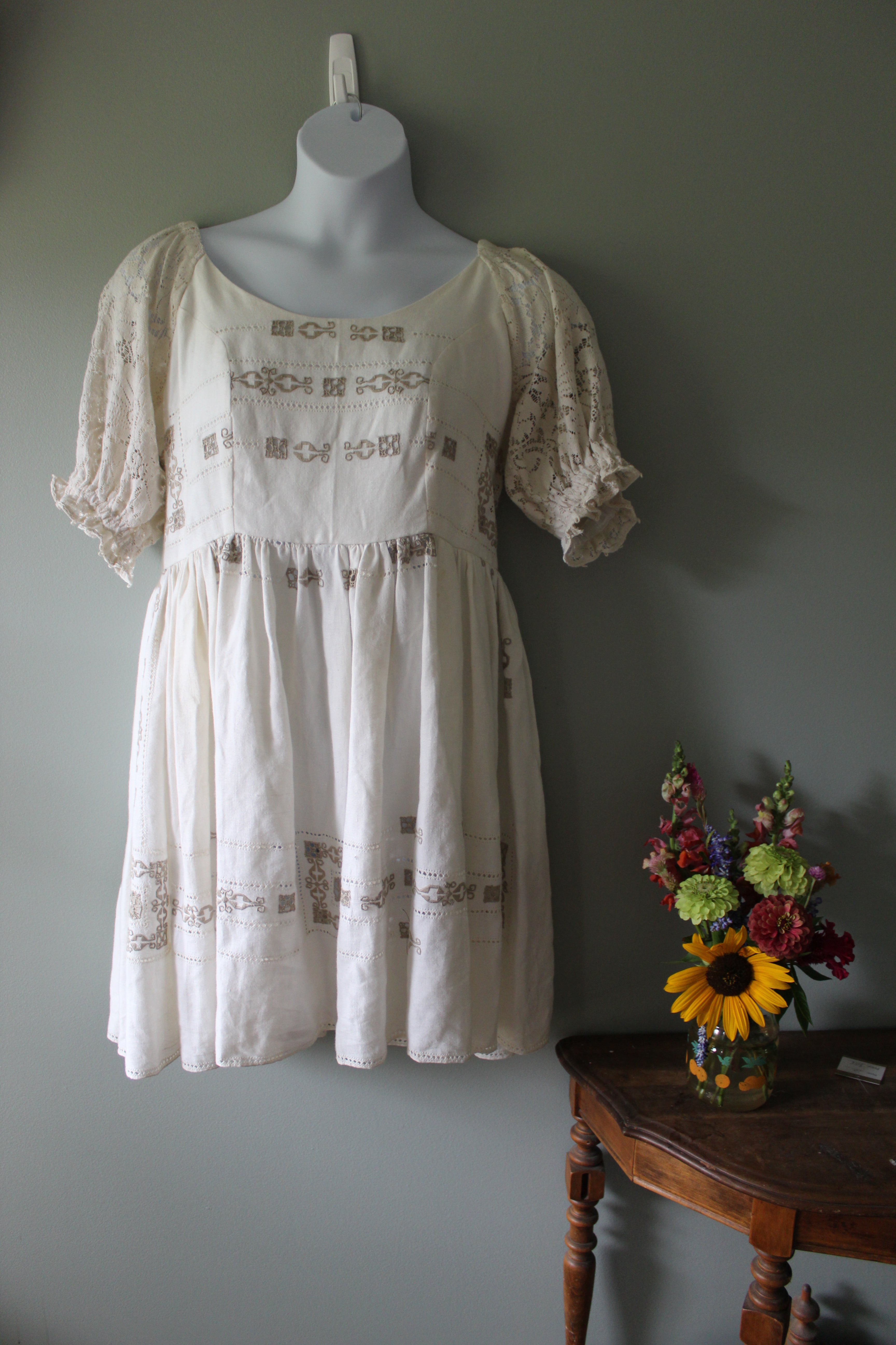 White dress with embroidery on a mannequin against a gray wall with flowers on a chair.