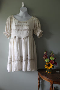 White dress with embroidery on a mannequin against a gray wall with flowers on a chair.